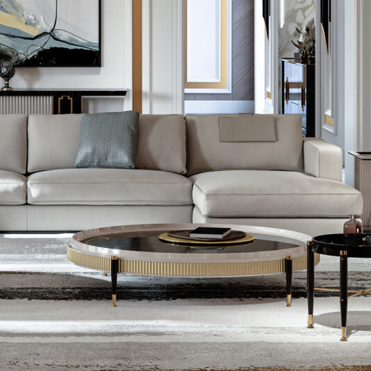 A modern living room showcasing the Large Chaise Art Deco Style Sofa in light gray, adorned with a blue-green throw pillow. In front of the sofa sits a round coffee table featuring a glass top and gold accents. The room maintains a neutral color palette, complemented by art and décor on the walls.