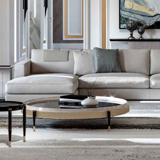 A modern living room featuring a high-end Art Deco style, with a Large Chaise Art Deco Style Sofa in light gray, adorned with blue and gray throw pillows. In front of the sofa, there is a round glass coffee table showcasing decorative items and books. The room boasts detailed wall paneling and a neutral color palette.