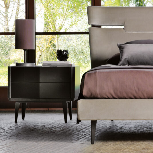 Modern Bedside Cabinet With Two Drawers Modern bedroom featuring a beige upholstered bed with purple bedding. A dark wood nightstand beside the bed holds a lamp and decorative items. Large windows in the background reveal a lush green landscape, complemented by the Modern Bedside Cabinet With Two Drawers for extra storage.