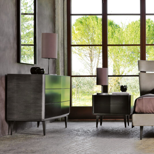 Modern Bedside Cabinet With Two Drawers A modern bedroom is adorned with a large window offering a view of lush green trees, two purple lamps on a dark wooden chest of drawers and bedside table, a mirror above the chest of drawers, and a bed dressed in neutral-colored linens. The space also features the Modern Bedside Cabinet With Two Drawers, concrete-textured flooring, and walls painted grey.