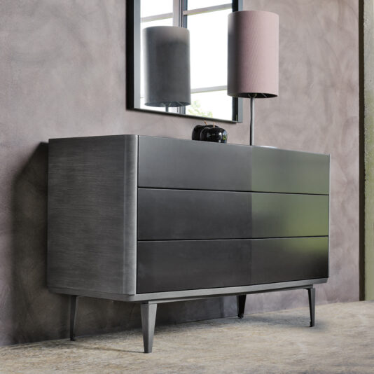 A sleek, modern chest of drawers in gray with three spacious compartments from the "Modern Chest Of Drawers" collection stands against a textured wall. A matching gray table lamp with a pink shade and a small black clock adorn the top. Above the dresser, a reflective window adds light to the room. The floor is finished in concrete.