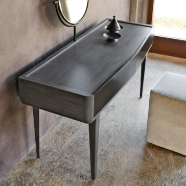 Modern Dressing Table With Mirror The Modern Dressing Table With Mirror, characterized by sleek, clean lines, is positioned against a textured wall. This wooden table features a small, decorative metallic incense burner on its surface. Above it hangs a round mirror, and to the right sits a square, light-colored ottoman.