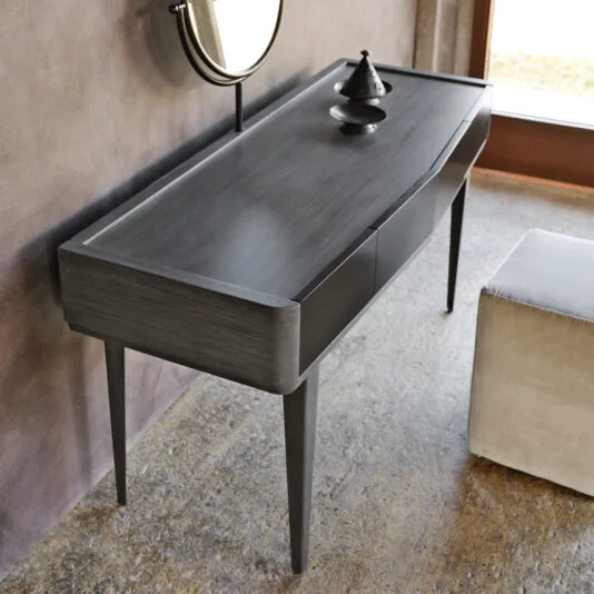 The Modern Dressing Table With Mirror, characterized by sleek, clean lines, is positioned against a textured wall. This wooden table features a small, decorative metallic incense burner on its surface. Above it hangs a round mirror, and to the right sits a square, light-colored ottoman.