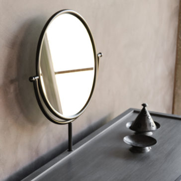 Modern Dressing Table With Mirror A Modern Dressing Table With Mirror stands near a window with crossbars, reflecting light off its black-framed, round surface. In the foreground rests a small metallic, conical container with a lid on a saucer. The wall in the background features a muted, textured shade.
