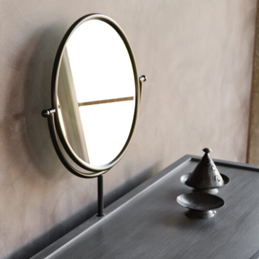 A Modern Dressing Table With Mirror stands near a window with crossbars, reflecting light off its black-framed, round surface. In the foreground rests a small metallic, conical container with a lid on a saucer. The wall in the background features a muted, textured shade.