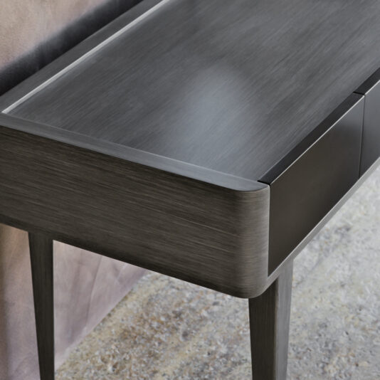 The Modern Dressing Table With Mirror boasts a sleek, contemporary design featuring curved edges and a matte finish. This wooden console stands gracefully on four slim legs and includes a drawer with a minimalist, handle-less design. The background features a textured wall and stone floor.