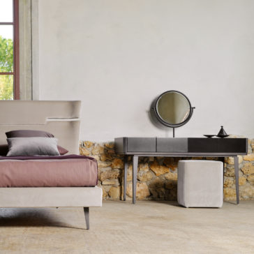 Modern Dressing Table With Mirror A minimalist bedroom features a beige bed adorned with a brown and gray bedspread. Beside the bed is the Modern Dressing Table With Mirror, complemented by a small gray ottoman. The room boasts a stone accent wall and a window overlooking vibrant greenery.