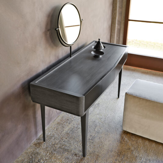 The Modern Dressing Table With Mirror, characterized by its sleek, modern design and tapered legs, includes a round mirror mounted on the wall above it. A small decorative item adorns the table's surface. Beside the table, a cushioned stool upholstered in neutral-toned fabric offers comfortable seating. The area is illuminated by natural light streaming in through a large window.