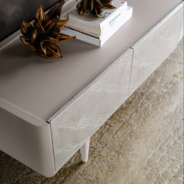 The Modern Low Sideboard With Drawers features smooth, light-colored surfaces and subtly textured fronts. Decorative metallic artichokes and a stack of books adorn the top. The sideboard stands on four slender legs against a textured, neutral-toned floor.