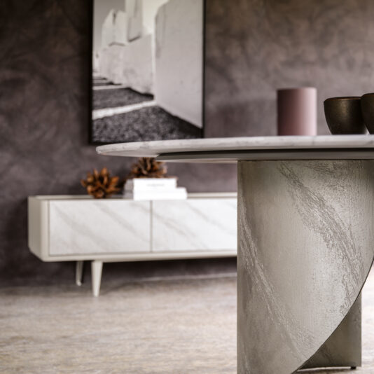 A contemporary living room features a round marble table adorned with a ceramic cup and vase. Against the back wall stands the Modern Low Sideboard With Drawers, above which hangs a black-and-white photograph. The room is characterized by its neutral color palette and textured walls.