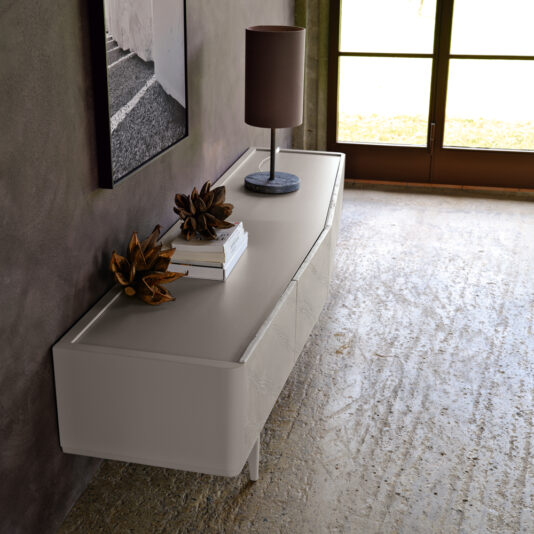 A contemporary living room showcases a Modern Low Sideboard With Drawers, elegantly embellished with decorative objects, books, and a table lamp featuring a gray base and brown shade. Above the sideboard, a framed black-and-white photograph is displayed on the wall as natural light pours in from nearby windows.