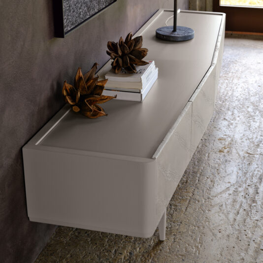 A Modern Low Sideboard With Drawers, showcasing a sleek, light-colored surface and a textured front, is placed next to a rough-textured wall. The sideboard holds a small stack of books, a contemporary lamp, and two decorative sculptures resembling dried flowers.