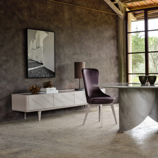 A modern interior showcasing the sleek Modern Low Sideboard With Drawers, embellished with tasteful decor items, a framed abstract artwork on the wall, a contemporary round table with a textured base, and an elegant chair featuring dark cushions. Natural light floods in through a large window, illuminating the entire space.