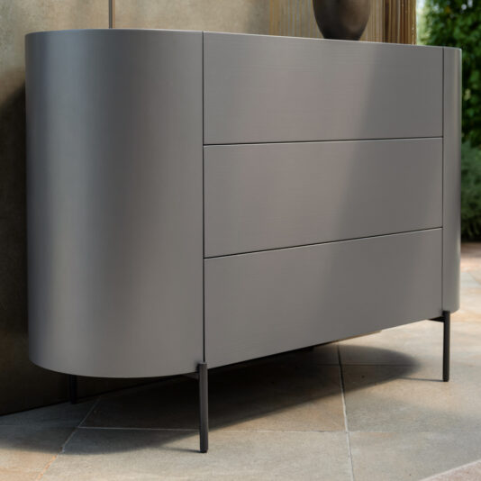 Modern Minimal Curved Chest Of Drawers The Modern Minimal Curved Chest Of Drawers, exhibiting a sleek and modern gray finish with three elegantly curved drawers, is placed in a contemporary setting. This sophisticated dresser features rounded edges and stands on minimalist legs. The backdrop includes a concrete wall accompanied by some greenery, evoking an indoor-outdoor ambiance.