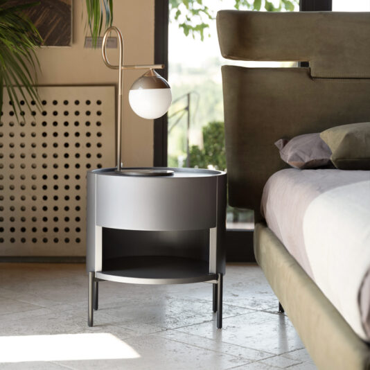 Modern Minimal Round Bedside Cabinet A modern bedroom showcases the Modern Minimal Round Bedside Cabinet, which boasts a sleek design with a built-in lamp. The cabinet has a matte black finish and features a single open shelf beneath it. Adjacent to the cabinet is a bed with green upholstery, while natural sunlight streams in through a window in the background.