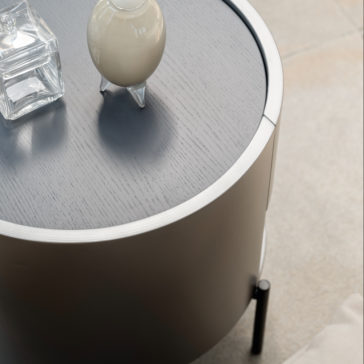 Modern Minimal Round Bedside Cabinet The Modern Minimal Round Bedside Cabinet, finished in a sleek gray, features a stylish and modern design. It is topped with a clear square glass container and a small white vase-like object. Placed on a beige tile floor, this arrangement enhances the minimalist and clean aesthetic of the space.