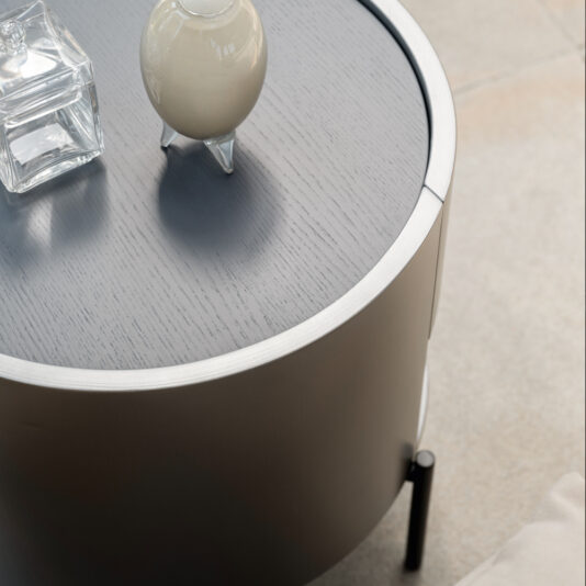 Modern Minimal Round Bedside Cabinet The Modern Minimal Round Bedside Cabinet, finished in a sleek gray, features a stylish and modern design. It is topped with a clear square glass container and a small white vase-like object. Placed on a beige tile floor, this arrangement enhances the minimalist and clean aesthetic of the space.