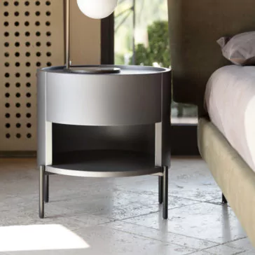 Modern Minimal Round Bedside Cabinet The Modern Minimal Round Bedside Cabinet in gray, featuring a drawer and an open shelf below, stands on sleek legs beside a bed with light bedding. A spherical lamp is positioned on the cabinet, and in the background, you can see a perforated screen and outdoor greenery.