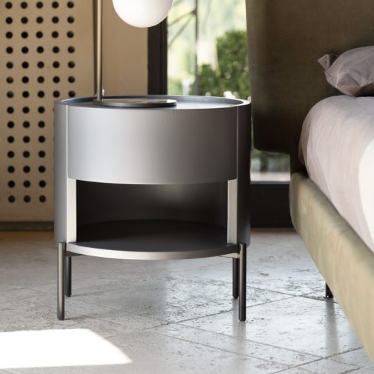 The Modern Minimal Round Bedside Cabinet in gray, featuring a drawer and an open shelf below, stands on sleek legs beside a bed with light bedding. A spherical lamp is positioned on the cabinet, and in the background, you can see a perforated screen and outdoor greenery.