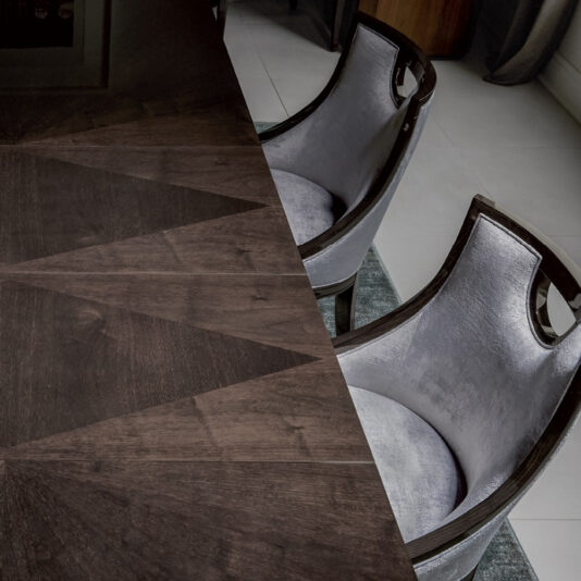 Modern Walnut Luxury Dining Chair A sleek, modern dining table with a geometric wooden pattern is accompanied by two Modern Walnut Luxury Dining Chairs, featuring luxurious walnut seats and dark wooden frames. The light gray floor and part of a dark wooden cabinet are visible in the background, enhancing the contemporary interior design.