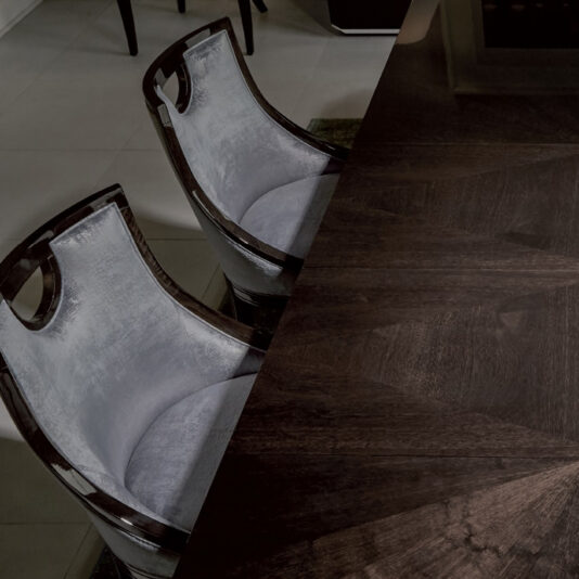 Modern Walnut Luxury Dining Chair A close-up view of two Modern Walnut Luxury Dining Chairs with wooden frames tucked under a dark wooden table. The chairs feature a curved backrest design and are positioned on a light-tiled floor. Part of the table's surface is visible, revealing its rich, dark wood grain.