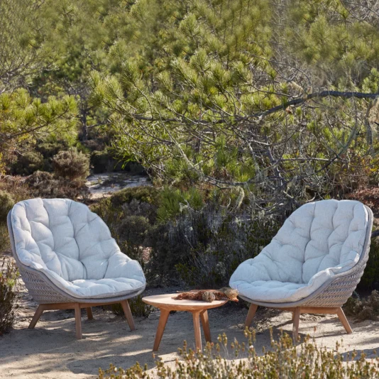 Contemporary Outdoor Woven Armchair Two Contemporary Outdoor Woven Armchairs surround a small round table in a natural setting of trees and shrubs, evoking a peaceful, sunny day focused on nature and relaxation.