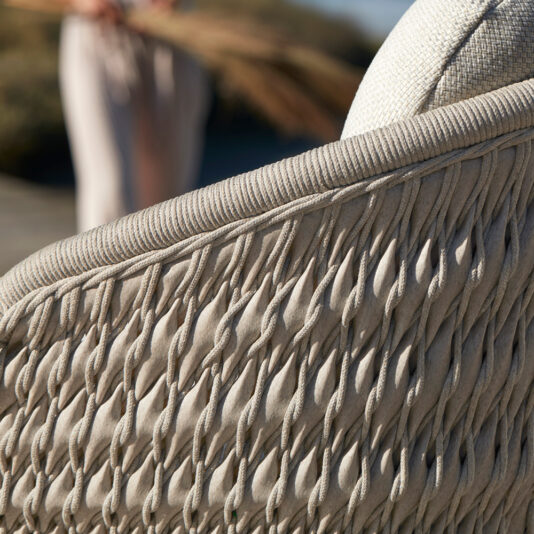Contemporary Outdoor Woven Armchair A close-up of the Contemporary Outdoor Woven Armchair featuring a textured, intricately braided design with a neutral-colored cushion, highlighted beautifully in the sunlight. In the blurred background, an individual is standing outdoors while holding an object.