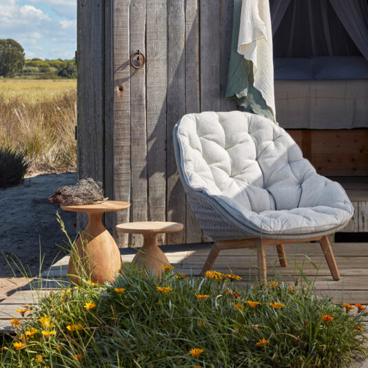 Contemporary Outdoor Woven Armchair A cozy outdoor setting features a Contemporary Outdoor Woven Armchair on a wooden deck, flanked by two rustic wooden side tables. A wooden cabin with an open door and hanging linens in the background. Yellow and orange flowers grow in the foreground.