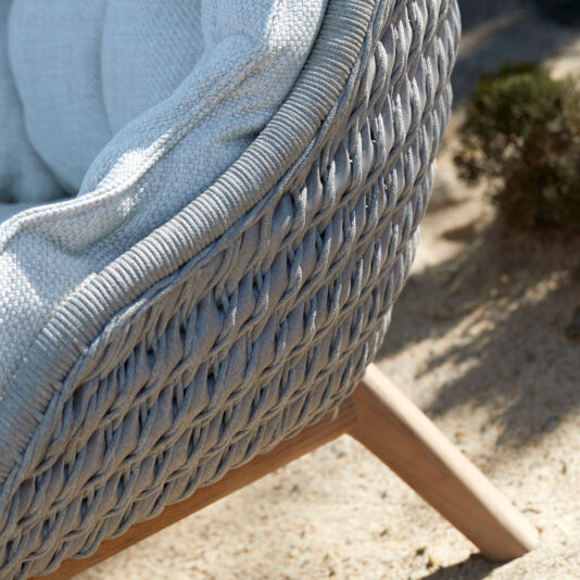 Contemporary Outdoor Woven Armchair Close-up of the Contemporary Outdoor Woven Armchair with light blue cushions. The chair features a textured, braided design and sturdy wooden legs. In the background, sandy ground and small greenery suggest a serene outdoor setting.