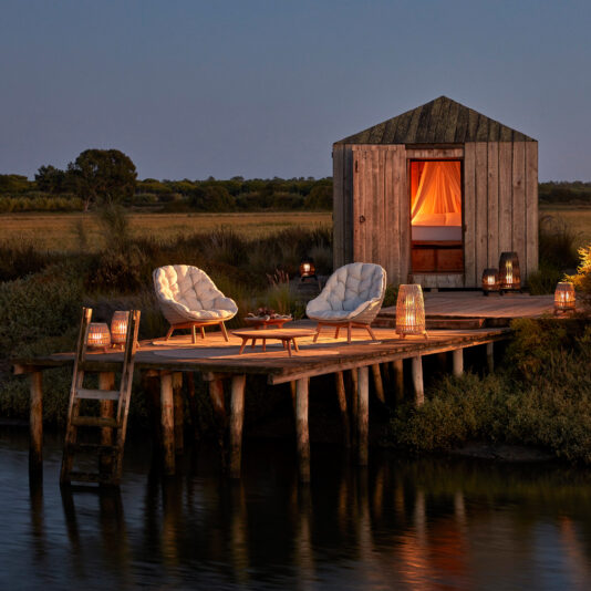 Contemporary Outdoor Woven Dining Chair A serene evening scene of a wooden deck over water, featuring Contemporary Outdoor Woven Dining Chairs and lanterns creating a cozy atmosphere. In the background, a small rustic cabin with a warmly lit interior sits amidst lush greenery.