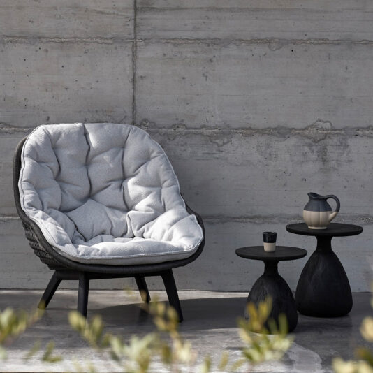 Contemporary Outdoor Woven Armchair A Contemporary Outdoor Woven Armchair is cozily situated against a concrete wall, flanked by two small black side tables. One table supports a cup, while the other holds a teapot. Out-of-focus plants in the foreground frame the scene, enhancing the relaxing outdoor ambiance.