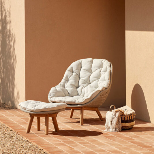 A Contemporary Outdoor Woven Armchair, paired with a matching ottoman, enhances the terracotta tile patio that stands against the stucco wall. Adjacent to the chair, a woven basket holding a cozy blanket completes the inviting and stylish outdoor seating area.