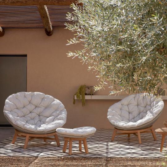 Two cozy, round, cushion-filled chairs and a matching ottoman are arranged on a patterned tiled patio. An olive tree partially shades the seating area while the Contemporary Outdoor Woven Daybed adds elegance. A rustic beam roof and a beige wall with a small shelf in the background complete the serene setting.