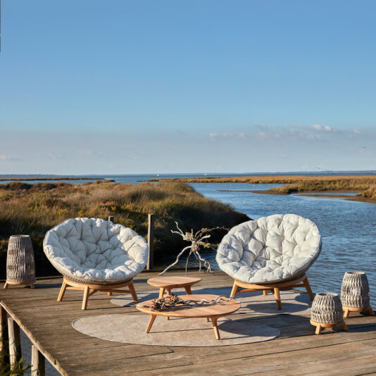 Contemporary Outdoor Woven Daybed In a serene outdoor seating area overlooking a calm water body and grassy landscape, you'll find a Contemporary Outdoor Woven Daybed accompanied by two cushioned, round lounge chairs, a low wooden coffee table, and several small stools. The clear sky enhances the peaceful ambiance.