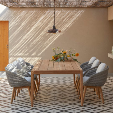 A stylish outdoor dining area featuring a long wooden table surrounded by six Contemporary Outdoor Woven Dining Chairs. The space is decorated with a floral centerpiece and shaded by a textured canopy casting intricate shadows. The floor features a geometric tiled pattern.