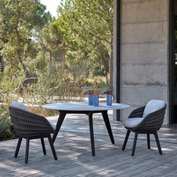 A sunny outdoor patio showcases a wooden deck with a round table and two Contemporary Outdoor Woven Dining Chairs. The table, adorned with two blue vases filled with decorative sticks, is surrounded by lush greenery and trees next to a concrete building wall.