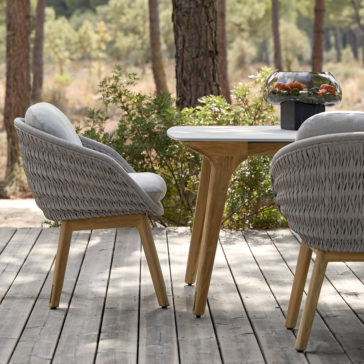 An outdoor wooden deck features a table and two Contemporary Outdoor Woven Dining Chairs. The table has light-colored wooden legs and a gray surface, while the chairs boast light gray cushioning in a woven design. On the table rests a glass bowl filled with fruits, and trees are visible in the background.