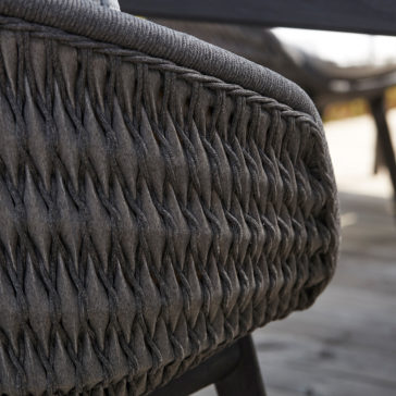 Close-up image of the Contemporary Outdoor Woven Dining Chair. The chair showcases tightly woven dark fibers that form a patterned, intricate design. A slightly blurred wooden deck in the background suggests an outdoor setting.