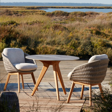 A wooden patio table and two Contemporary Outdoor Woven Dining Chairs with cushions are placed on a deck overlooking grasslands and a distant body of water. The scene exudes a peaceful, natural ambiance suitable for outdoor relaxation.