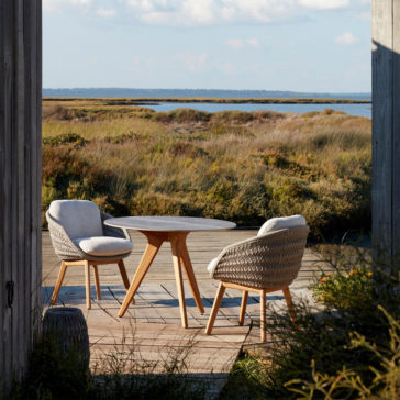 The outdoor seating area features two Contemporary Outdoor Woven Dining Chairs and a round table, all set on a wooden deck that overlooks scenic marshland. Tall grasses and shrubs surround the deck, with a tranquil body of water and a hazy, distant horizon under a partly cloudy sky.