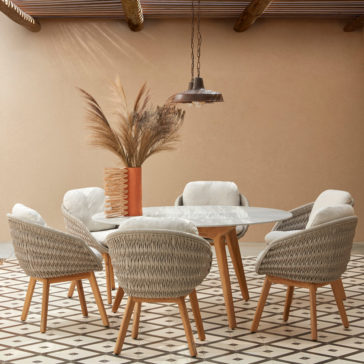 A stylish dining area featuring a round white table surrounded by six Contemporary Outdoor Woven Dining Chairs with light cushions. A tall vase filled with dried pampas grass sits in the center. The space has beige walls, a patterned tile floor, and a pendant light hanging above.