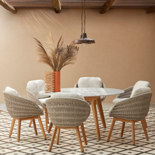A stylish dining area featuring a round white table surrounded by six Contemporary Outdoor Woven Dining Chairs with light cushions. A tall vase filled with dried pampas grass sits in the center. The space has beige walls, a patterned tile floor, and a pendant light hanging above.