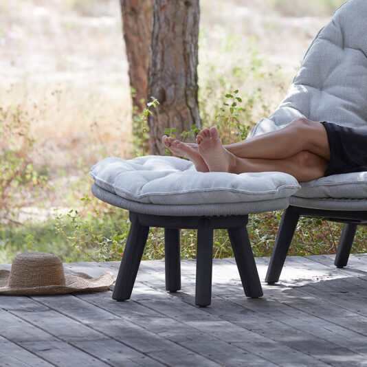 A person is unwinding with legs propped on a Contemporary Outdoor Woven Footstool on a wooden deck, surrounded by trees. Nearby, a straw hat lies on the ground, enhancing the tranquil, leisurely atmosphere.