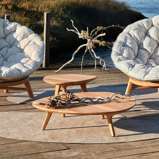 Two plush, cushioned chairs surround two Contemporary Teak Outdoor Coffee Tables on a wooden deck. Dried branches are arranged decoratively on the tables. In the background, there's a grassy area by the water.