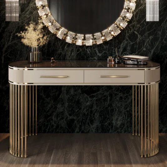 An Art Deco Style Ribbed Console Table with metallic accents is set against a dark marble wall. It features two drawers and is accessorized with an ornate round mirror, a vase of dried plants, and books on top.