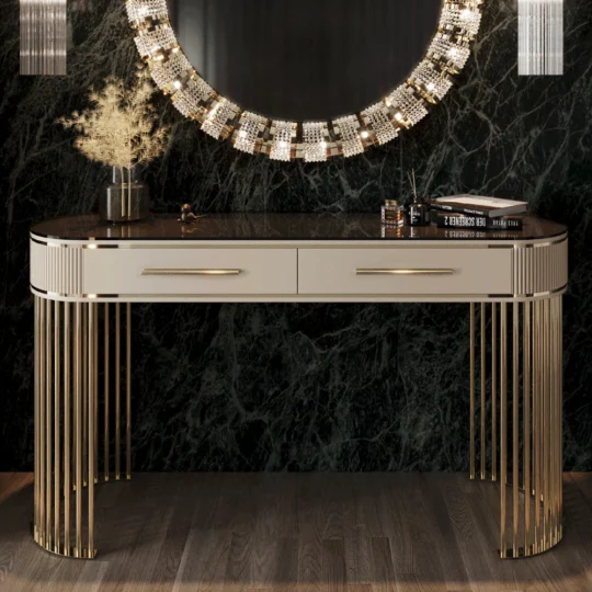 An Art Deco Style Ribbed Console Table with metallic accents is set against a dark marble wall. It features two drawers and is accessorized with an ornate round mirror, a vase of dried plants, and books on top.