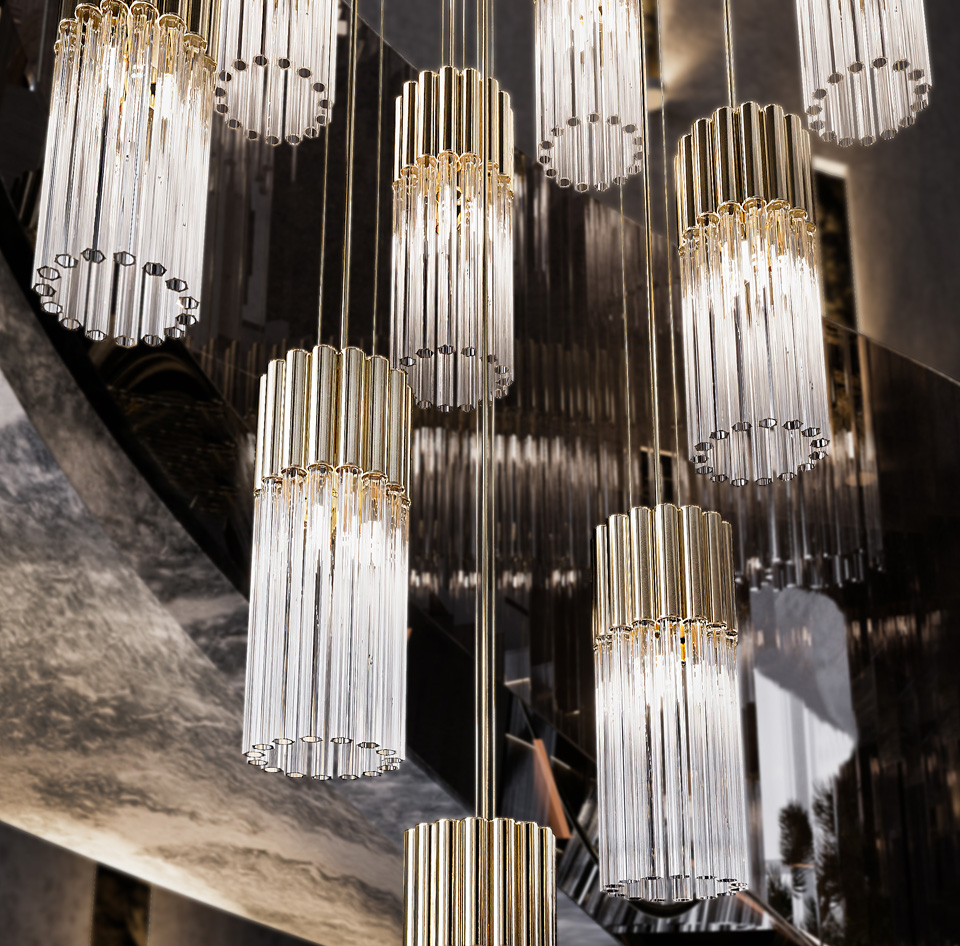 Contemporary Glass Tube Pendant Light A collection of Contemporary Glass Tube Pendant Lights featuring gold accents and vertical glass rods gracefully suspend from the ceiling. These elegant lights are artistically varied in height and arranged against a sophisticated, reflective background.
