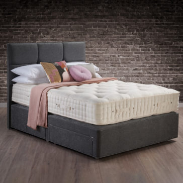 Hypnos Luxury Wool Origins 10 Mattress A large, well-constructed bed with a dark upholstered headboard and convenient under-bed storage drawers showcases the Hypnos Luxury Wool Origins 10 Mattress. The bed is adorned with multiple pillows, a folded pink blanket, and a textured throw in hues of pink, beige, and red. A brick wall serves as the backdrop.