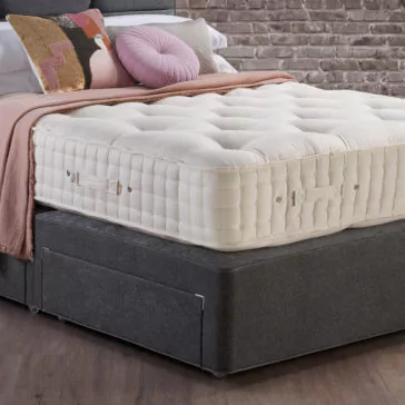 Hypnos Luxury Wool Origins 10 Mattress A bedroom scene showcasing a rich Hypnos Luxury Wool Origins 10 Mattress resting on a dark grey base. The bed is embellished with a light pink throw blanket and several decorative pillows, including a round pink one and a rectangular pillow with a geometric pattern. A brick wall provides the backdrop.