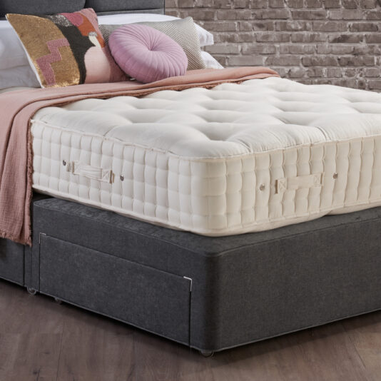 A bedroom scene showcasing a rich Hypnos Luxury Wool Origins 10 Mattress resting on a dark grey base. The bed is embellished with a light pink throw blanket and several decorative pillows, including a round pink one and a rectangular pillow with a geometric pattern. A brick wall provides the backdrop.